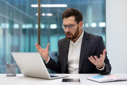 A worried young businessman sits at a desk in the office, looks at the laptop screen and spreads his arms in frustration