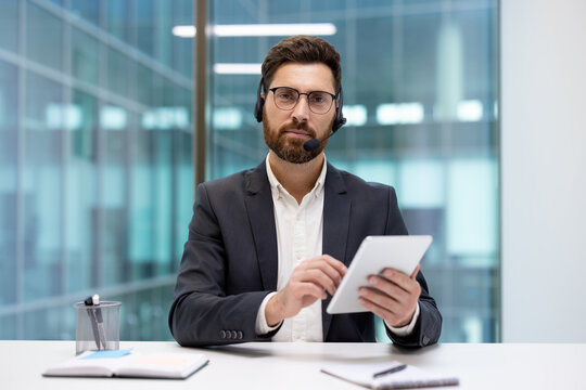 A portrait of a serious young man in a suit and glasses sitting at a desk in an office, holding a tablet and looking at the camera