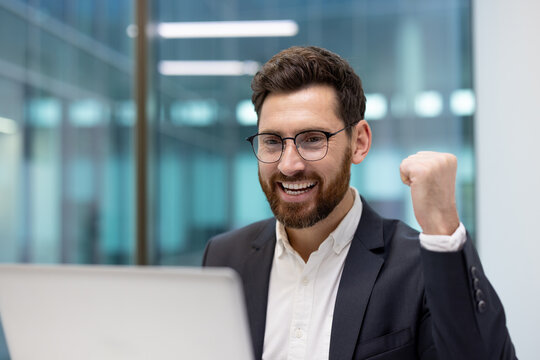 Close-up photo of a happy young male businessman sitting in the office, looking at the laptop screen and rejoicing in success