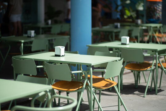 Sunlit outdoor dining area with pastel metal furniture, wooden chair seats and napkin dispensers beside a blue column, with selective focus highlighting the inviting sidewalk seating