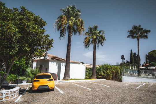 A golden hatchback anchors an asymmetric composition of green palms, white stucco and blue sky, while diagonal cobbled parking bays create depth in a vivid sunlit holiday setting
