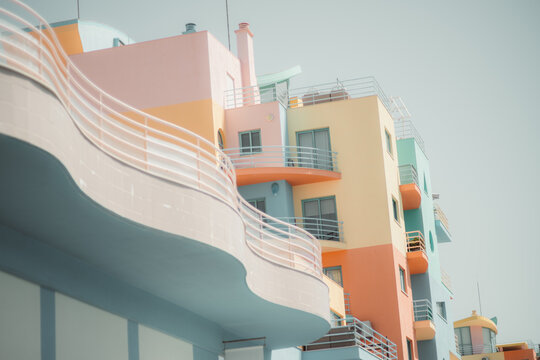 Peach, pink, yellow and aqua facades rise behind flowing white railings, with rounded terraces and geometric windows creating a playful architectural composition against a pale clear sky
