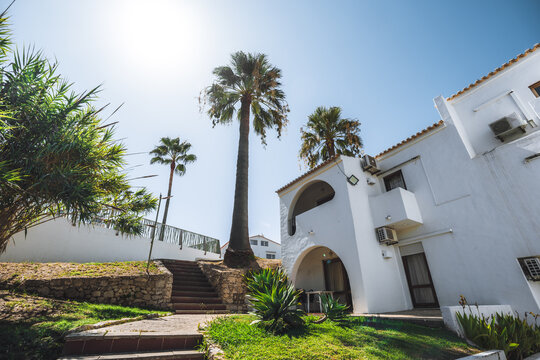 Bright stucco accommodation with arched balconies and tiled roofs overlooks a landscaped garden, where stone steps climb past tall tropical trees beneath clear blue skies and strong sunshine