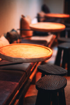 Round wooden tabletops, dark stools and cushioned bench seating glow in amber light inside a cozy restaurant, with shallow depth of field softening the background into warm atmospheric blur