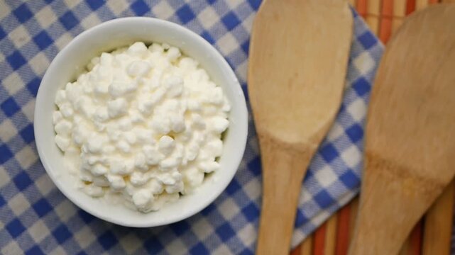 Top view of cottage cheese in white bowl with wooden spoons on checkered cloth dynamic motion video