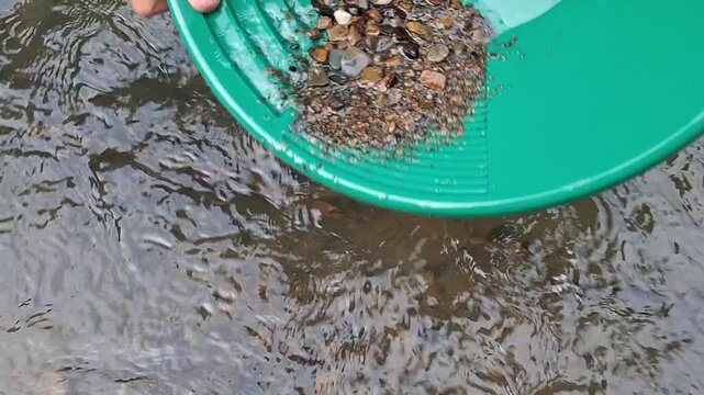 Slow motion close-up footage of a gold prospector carefully removing large pebbles and stones from a green plastic gold pan. The hand gently pushes and discards coarse gravel 