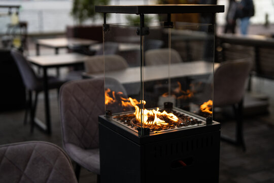 Street cozy terrace with comfortable seating places and gas heater in glass box for guests, modern restaurant outdoor patio with couches, tables and fireplace inside glass cabinet, dining lounge zone.