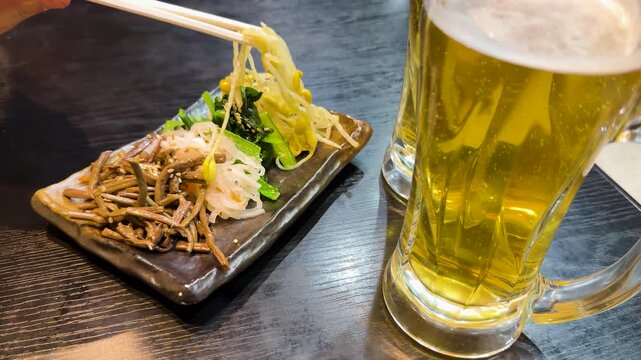 A close up shot of a person picking bean sprouts with chopsticks from a plate of assorted namul with two mugs of beer on a black table.