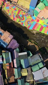 Top-down drone view over colorful Jodipan Village in Malang, Indonesia, showing painted rooftops, riverside murals, bridge and river in a vibrant urban neighborhood.