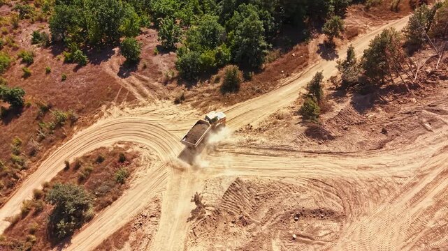 A dump truck drives on a dirt road in a construction area. The vehicle stirs up dust as it travels. Trees line the pathway under clear skies.