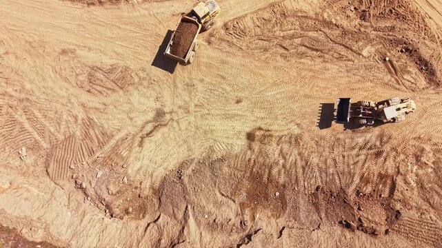 Two pieces of construction equipment are on a dirt site moving soil and materials under clear skies during the daytime. Tire tracks are visible in the dirt.