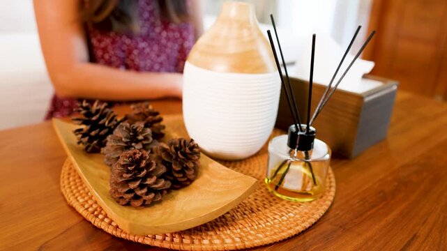 Woman sitting behind table with wooden aroma diffuser and pine cones decoration in modern living room.