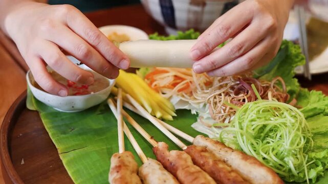 Person preparing Vietnamese spring rolls with pork skewers and fresh herbs on banana leaf.