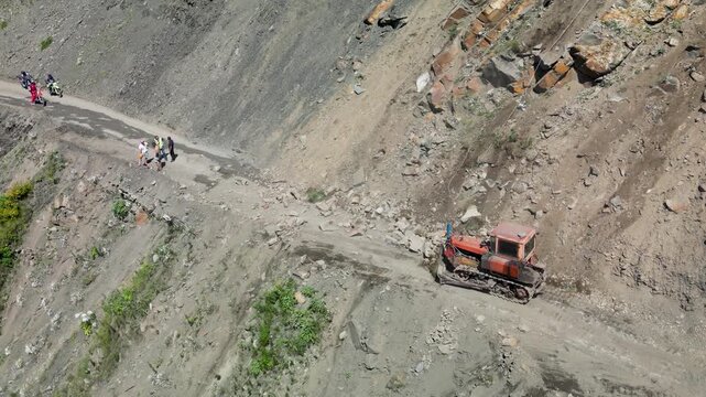 Aerial view of an excavator loading and removing rock debris from a dirt road at the base of a steep mountain slope.