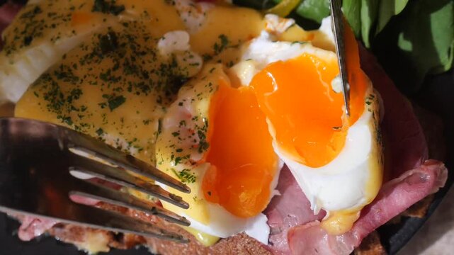 Close up cinematic footage of a knife and fork cutting into a perfectly poached egg, revealing a runny yolk on a delicious breakfast toast