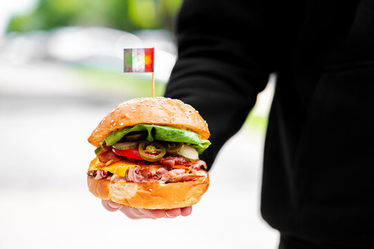A person's hand holding a juicy burger with pulled meat, cheese, jalape&ntilde;os, and lettuce, topped with a small Italian flag toothpick against a blurred background.