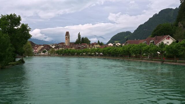 Turquoise Aare River flows past traditional buildings and greenery in Interlaken, Switzerland, with church tower and Swiss Alps under cloudy sky. 4K UHD video.
