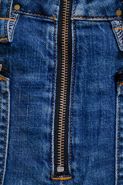Close-up of blue denim jeans front fly featuring a metal zipper with black leather pull tab, rivets, zippered side pockets, orange contrast stitching, and distressed fabric texture.