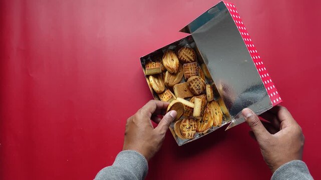 Close up slow motion video of hand taking a cookie from a box full of various delicious cookies on a red background