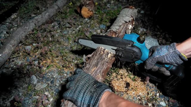 Person wearing protective gloves cutting a fallen birch tree log with a small cordless electric chainsaw, with sawdust flying around during nighttime yard work or land clearing activity