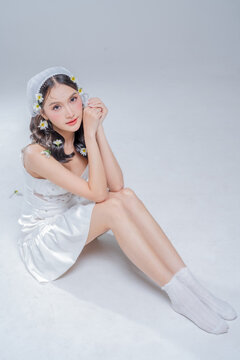 Graceful young woman sitting on studio floor in white silk dress, expressing romantic elegance, pristine beauty, poetic introspection, gentle vulnerability, serene purity, and modern bridal charm.