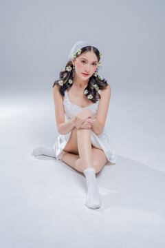 Graceful young woman sitting on studio floor in white silk dress, expressing romantic elegance, pristine beauty, poetic introspection, gentle vulnerability, serene purity, and modern bridal charm.