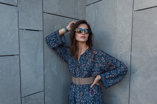 Stylish young woman in a patterned blue belted dress and sunglasses poses with hand in hair against a grey textured concrete wall. Modern urban fashion.