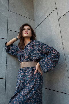 Low-angle portrait of an elegant brunette woman wearing a floral patterned blue dress with a wide belt, posing against a modern grey stone wall outdoors. Fashionable urban look.