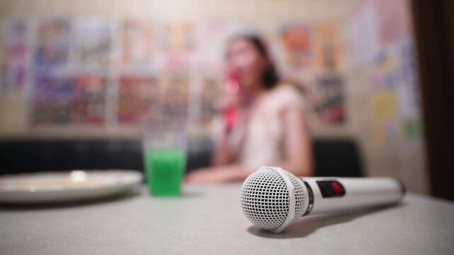 手前のマイクと奥に歌う女性がいるぼやけたカラオケルームの風景