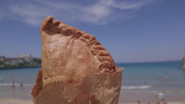 POV of a hand holding a Cornish Pasty above the beach in Newquay, Cornwall, England on a hot sunny June day.