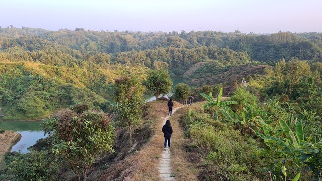 Two people hiking on a narrow dirt path along a ridge surrounded by lush green hills and a scenic lake under a soft sky