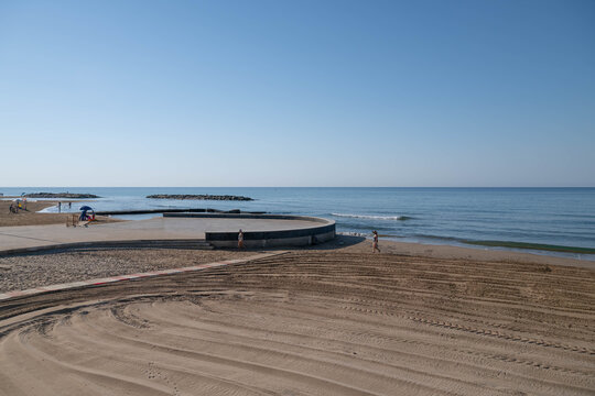 Mediterranean Sea and sandy beach under a clear blue sky, with gentle waves and people walking along the shore.