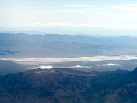 Aerial view of desert mountains overlooking a dry lakebed and glowing solar thermal power plants with mirror fields, Mojave Desert, California