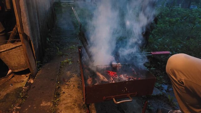 Delicious pieces of meat cooking on a hot charcoal brazier grill during a backyard barbecue party at dusk, with smoke billowing from the glowing embers as a man tends to the food