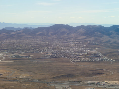 Aerial view of suburban desert sprawl in Nevada backed by mountain ranges, with a highway cutting through open scrubland