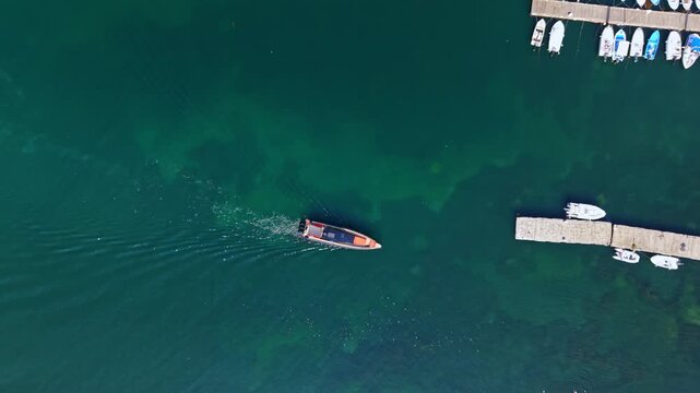 A boat travels over calm water close to a dock area filled with several boats. The scene shows clear blue water and sunny weather.