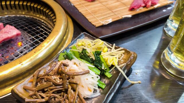 Japanese wagyu beef grilling on charcoal barbecue with namul appetizers and beer on wooden table.