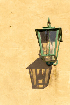 A green metal wall lantern with glazed sides that casts a distinct, geometric shadow onto the light, textured fa&ccedil;ade.
