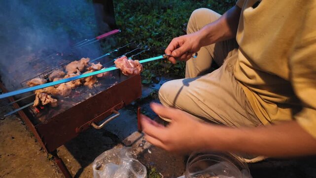 Unrecognizable man stringing fresh raw pork onto metal skewers for a shashlik barbecue at night, with other skewers already cooking over hot charcoal embers on a portable grill