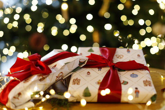 Christmas wrapped gifts tied with red ribbons on a wooden table illuminated by fairy lights, festive concept of gift giving. Holiday present wrapping, cozy Christmas Eve celebration.
