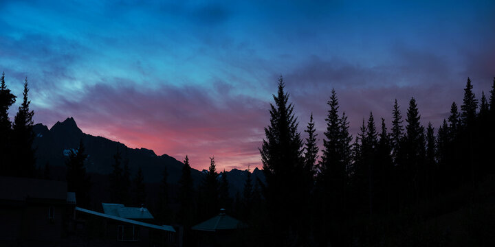 Sunrise panorama over mountain village and pine tree forest with pink clouds in the blue sky and mountain peaks in silhouette