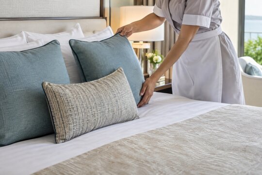 Professional housekeeper fluffing premium linen pillows on luxury hotel bed with neutral-toned decor, serene coastal view, pristine cleanliness
