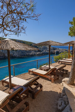 Serene Greek cove cabana lounger straw parasol crystal turquoise sea rugged cliffs Mediterranean summer. Peaceful summer vacation travel scene. Luxury relax wellness secluded coastal getaway