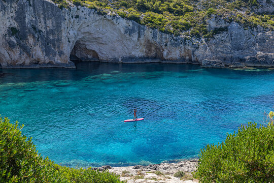 Paddleboarder exploring turquoise Greek lagoon dramatic limestone cliffs outdoor water sport summer adventure. Peaceful summer travel scene nature. Active recreation paddleboard coastal background