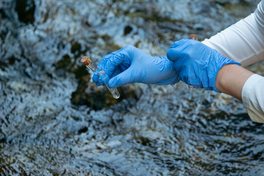 person in blue gloves holding glass tube with water sample outdoors testing for escherichia coli and gastrointestinal disease outbreak risk with copy space for banner design