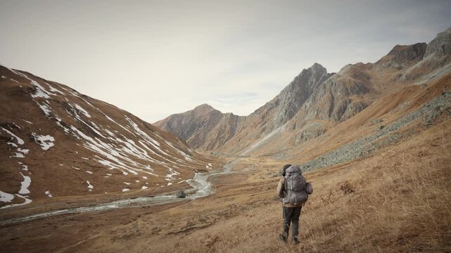 Trekker with heavy backpack walking away from camera into a vast golden Himalayan valley toward snow capped peaks in far west Nepal