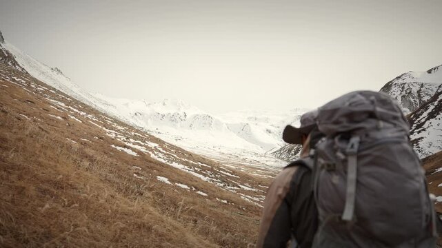 Trekker with heavy backpack walking away from camera in hazy golden valley slope descending toward distant snow covered Himalayan mountains under a soft muted sky in remote far west Nepal