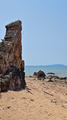 A tall weathered coastal rock formation stands beside a sandy, pebble-strewn beach overlooking calm turquoise water and distant islands under a clear blue sky. © อนุรักษ์ บุญมี