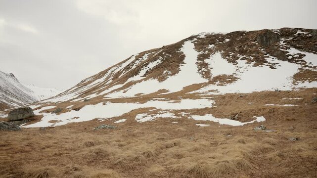 Empty golden valley meadow with scattered snow patches leading up toward a rounded snow dusted ridge under soft overcast Himalayan sky