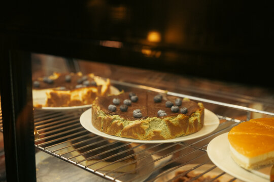 Freshly baked cheesecake cooling on wire rack inside warm bakery oven, with golden crust, blueberry topping, and orange cake nearby creating inviting dessert display